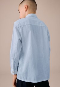 Lyseblå lang�ærmet skjorte lavet af et tekstureret stof. Har en standardkrave, knaplukninger ved ærmerne og en løs pasform.