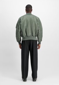 Man die met zijn rug naar de camera staat, gekleed in een groene bomberjack, losse zwarte broek en zwarte schoenen, tegen een effen witte achtergrond.