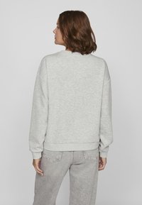 Mujer con cabello castaño hasta los hombros, vestida con un sudadera gris claro y unos jeans gris claro, de espaldas contra un fondo blanco liso.