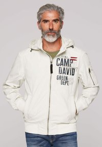 Light white jacket with a hood, featuring dark printed text, zippered pockets, and elastic cuffs. Worn over a green shirt and jeans.