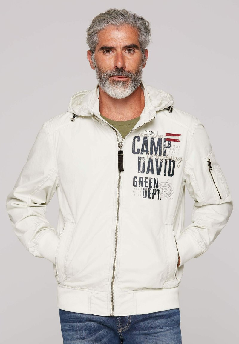 Light white jacket with a hood, featuring dark printed text, zippered pockets, and elastic cuffs. Worn over a green shirt and jeans.