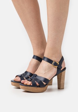 Navy leren sandalen met plateauzool, gevlochten bandjes, verstelbare enkelband met gouden gesp en een houten hak met textuur.