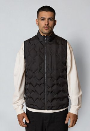 Hombre con chaleco acolchado negro con cremallera sobre una camisa blanca de manga larga, de pie frente a un fondo liso de color gris claro.