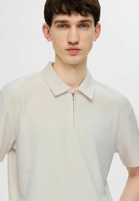 Světle béžové polo tričko s polovičním zipem v přední části, s texturou v jemném ribu a klasickým límcem. Materiál je hladký a lehký.
