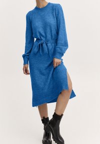 Robe en tricot bleu avec des manches longues, ceinture à la taille, poignets côtelés et fente sur le côté. Associée à des bottines noires. Tissu lisse et texturé.