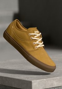 Yellow suede sneaker with white laces, rounded toe, and a flat brown rubber sole. Features branding on the tongue.