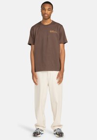 Camiseta de algodón marrón con mangas cortas que presenta un logo delantero en dorado. Combinada con pantalones holgados color crema y zapatillas negras.