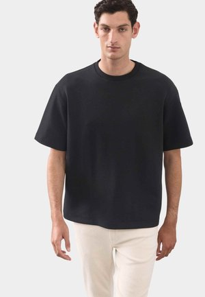 Mand iført en løstsiddende, sort kortærmet T-shirt og lyse bukser, stående mod en ensfarvet lys baggrund.