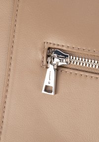 Beżowy materiał skórzany z srebrnym metalowym zamkiem błyskawicznym. Zamek posiada prostokątny uchwyt z wygrawerowanym wykrzyknikiem.