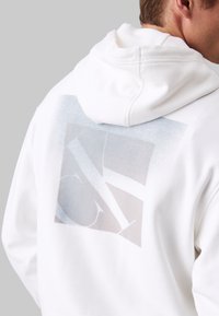 Mann in Weißem Hoodie mit abstraktem grau-blauen quadratischen Design auf dem Rücken, das geometrische Formen zeigt und eine Kapuze sichtbar ist.