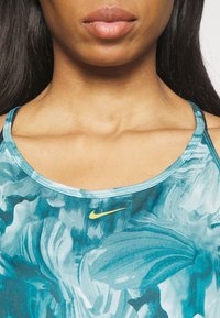 Débardeur de sport Nike avec imprimé floral bleu sarcelle. Comprend des bretelles fines, un col rond et un petit logo jaune sur la poitrine. Texture lisse.
