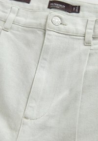 Hellbeige Jeans aus Denim in Nahaufnahme, die den Bund, die Vordertasche, den Metallknopf, die Gürtelschlaufen und das Etikett der N.PREMIUM-Kollektion in Größe 34R zeigen.