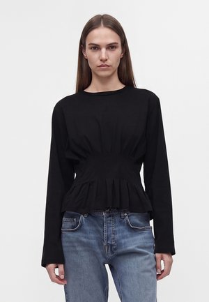 Femme aux longs cheveux bruns porte un haut péplum noir à manches longues froncé et un jean bleu, debout sur fond blanc.
