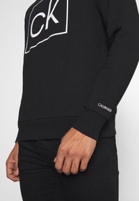 Sudadera negra de algodón, con un logo blanco de "CK" en un cuadrado, puños acanalados y una sutil etiqueta de "Calvin Klein" en la manga.