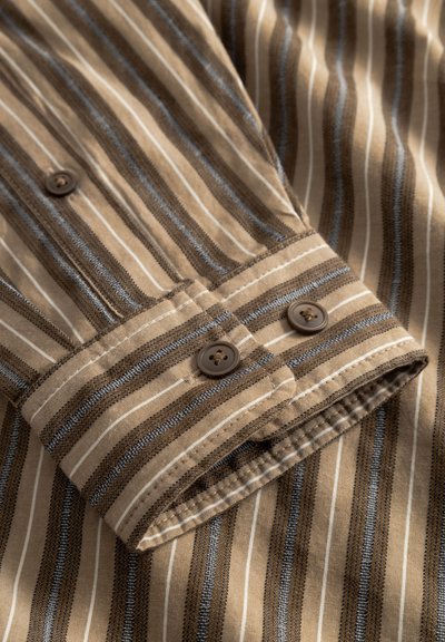 Zbliżenie na mankiet koszuli w beżowo-brązowe paski z dwoma brązowymi guzikami i szczegółowym przeszyciem na miękkiej tkaninie.