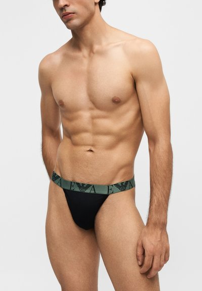 Homme torse nu portant un sous-vêtement noir avec une ceinture verte présentant de manière répétée la marque "EA" et un logo d'aigle.