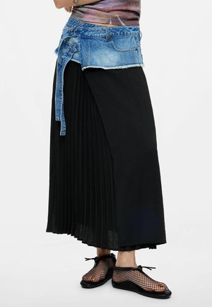 Person iført en sort plisseret nederdel med et påsat denimbælte og sorte netstrømpeballetsko, stående mod en ensfarvet baggrund.