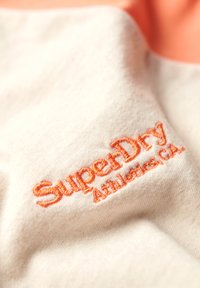 Cream fabric with orange embroidered "SuperDry" and smaller "Athletics.CA" text on a textured surface.