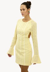 Vestido amarillo de encaje con mangas largas de campana, que presenta cortes en los lados y un patrón intrincado. Tejido texturizado con detalles de ribete vertical.