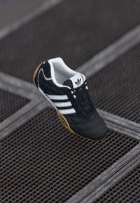 Tenisii negri Adidas cu dungi albe și talpă de cauciuc, așezați pe un podea din grilaj metalic.