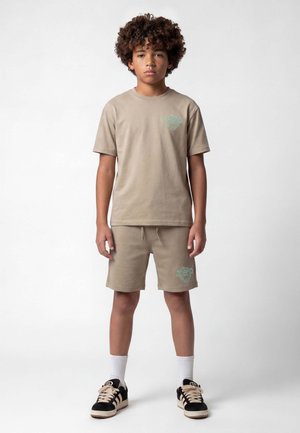 Boy with curly hair stands facing forward, wearing a beige t-shirt and shorts set with matching sneakers and white socks on a plain background.