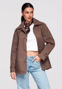 Kvinde iført brun knappet jakke, hvid kort top, blå jeans og mønstret halsklud, poserer med den ene hånd i lommen.