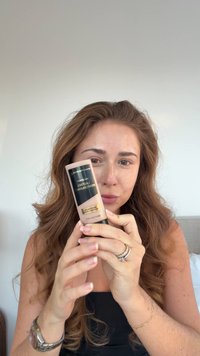 Max Factorin kasvopohjustus puoliläpinäkyvässä pullossa, jossa musta etiketti ja kultaiset yksityiskohdat. Sileä koostumus, pitkänomainen muoto ja ergonominen muotoilu.