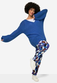 Blauwe gebreide oversized trui met V-hals, gecombineerd met gestreepte blauwe, bruine en witte leggings. Witte sneakers maken de outfit compleet.