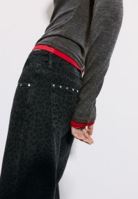 Črne jeans hlače iz denima z diskretno leopardsko vzorcem, srebrnimi dodatki, v kombinaciji s sivo majico z dolgimi rokavi, ki ima rdečo notranjo plast.