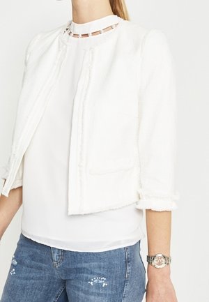 Kvinde iført en hvid tekstureret cropped jakke over en hvid bluse med perlekravedetaljer, kombineret med blå jeans og et sølvfarvet armbåndsur.