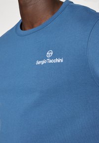 Camiseta de algodón azul con cuello redondo, diseño ajustado y logo bordado en blanco en el lado izquierdo del pecho. Textura suave y ligera.