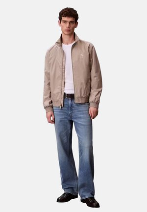 Giovane uomo che indossa una giacca bomber beige, una t-shirt bianca, jeans blu, cintura nera e scarpe nere, in piedi davanti a uno sfondo uniforme.