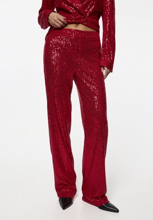 Femme portant un pantalon rouge brillant à sequins et un blazer assorti, associé à des chaussures noires à bouts pointus, debout devant un fond uni.