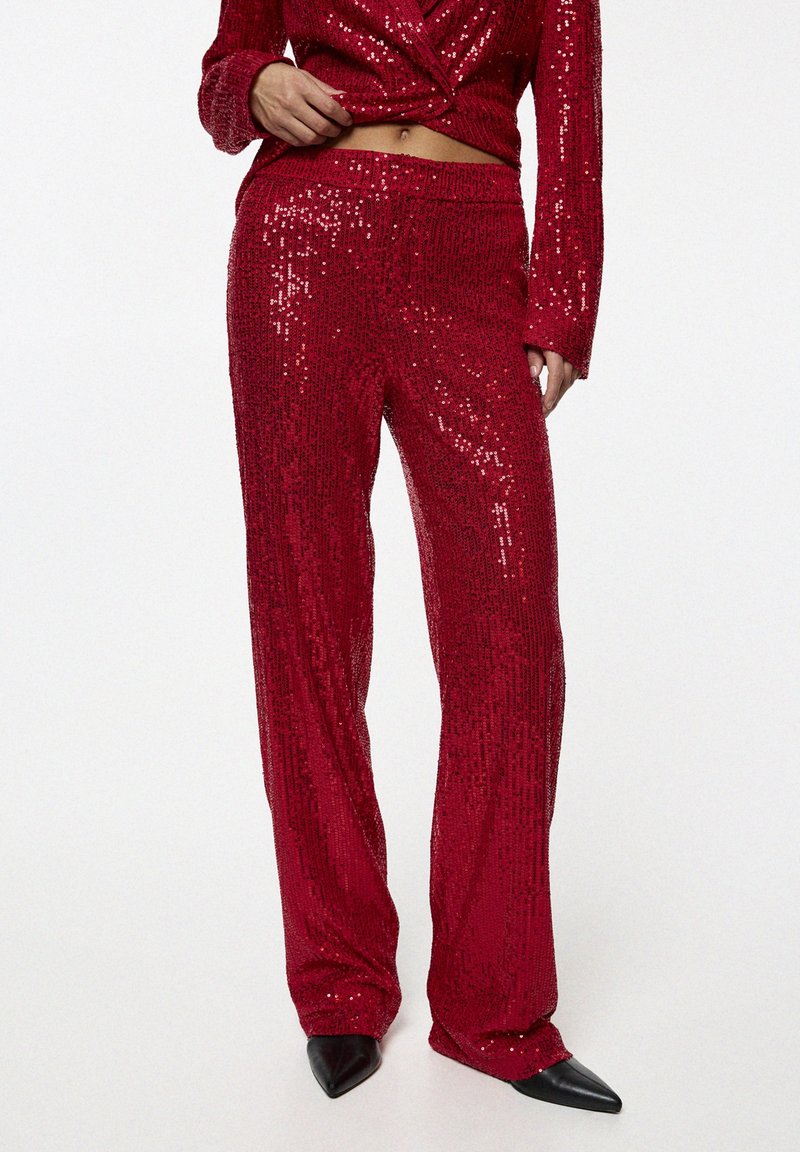 Femme portant un pantalon rouge brillant à sequins et un blazer assorti, associé à des chaussures noires à bouts pointus, debout devant un fond uni.