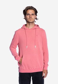 Roze hoodie van katoen, met een voorkrui pocket, koordjes en geribbelde manchetten. Het model draagt zwarte broek.