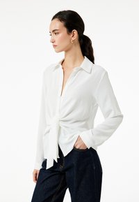 Witte overhem shirt met een geknoopte voorkant, gemaakt van textuurstof. Beschikt over een klassieke kraag en lange mouwen, gecombineerd met donkere denim.