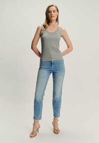 Mujer de pie con las manos detrás de la espalda, vistiendo una camiseta sin mangas gris, jeans de ligeros tonos azules, pendientes de aro dorados y sandalias de tacón beige.