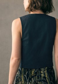 Schwarzes, ärmelloses Oberteil mit hohem Ausschnitt, strukturiertem Stoff, eng anliegendem Design, kombiniert mit einem gemusterten, fließenden Rock mit grünen und gelben Akzenten.