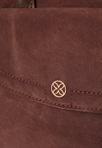 Smeđi antilop materijal s glatkom teksturom, šavom od šivanja i zlatnim kružnim logotipom s cvjetnim uzorkom.
