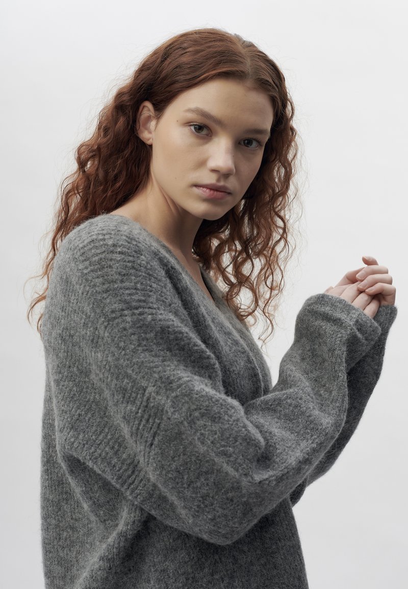 Grauer gestrickter Pullover mit tiefem V-Ausschnitt, langen, oversized Ärmeln und einer weichen Textur. Der Stoff wirkt flauschig und warm.