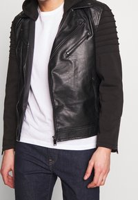 Giacca da motociclista nera in pelle con maniche in tessuto a coste, zip argento e cappuccio staccabile. Presenta una vestibilità slim e un design minimalista.