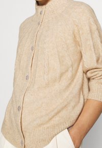 Cardigan en maille beige avec un col montant, présentant des motifs en tricot torsadé et des boutons décoratifs en pierres précieuses. Texture douce et ourlet côtelé.