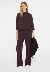 Violettes, strukturiertes Sweatshirt mit hohem Kragen, kombiniert mit weit geschnittenen passenden Hosen. Weiße, flache Sneakers und ein dunkler Strickschal sind enthalten.