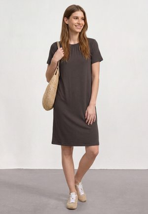 Frau trägt ein dunkelgraues Kleid mit kurzen Ärmeln, beige Sneakers und hält eine geflochtene Umhängetasche, steht auf einem grauen Boden vor einer weißen Wand.