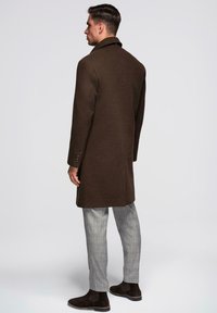 Ruskea villainen päällystakki, jossa on klassinen kaulus ja yksinäppäimiset kalvosimet, puettu ruudullisten housujen ja tummien nilkkureiden päälle, nähtynä takaa.