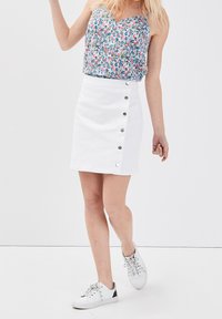Mini-jupe en denim blanc avec détails de boutons sur le côté, associée à un top sans manches à imprimé floral. Le modèle porte des baskets blanches. Design minimaliste.