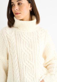 Cream-colored knitted sweater with a high ribbed collar, featuring cable knit patterns and a loose fit. Elaborate texture throughout.