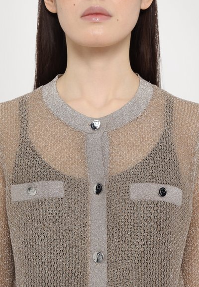 Beige, läpinäkyvä neulecardigan, jossa on hopeinen kaulus. Varustettu kahdella rintataskulla ja kiiltävillä, pyöreillä mustilla painikkeilla. Teksturoitu kangas, jossa on metallilankoja.