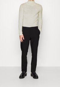 Krämfärgad polokrage med långa ärmar, kombinerad med svart skräddarsydda byxor. Svarta loafers har en kraftig sula, vilket ger en modern känsla till outfiten.