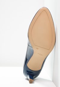 Marinblå patentläderhögklackad sko med spetsig tå och smal klack. Undersidan är beige med subtil märkning och storleksangivelser.